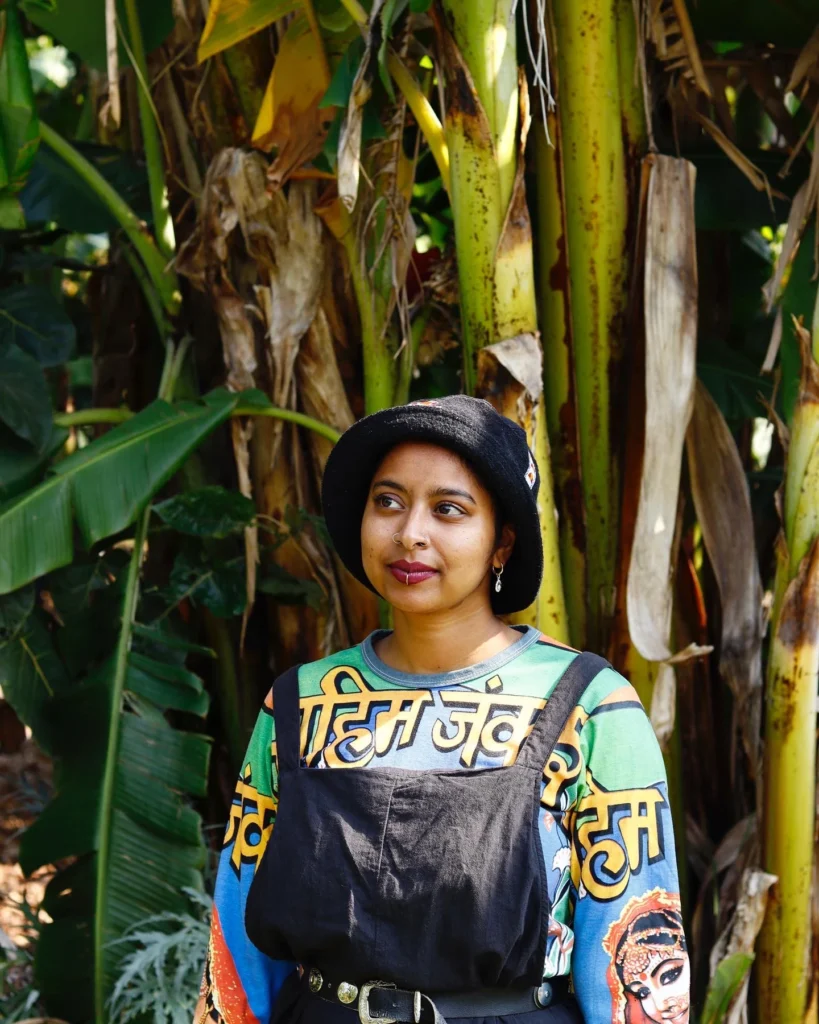 Youlendree Appasamy, a brown-skinned femme stands in front of banana leaf trees, wearing a black pair of dungarees over a top with Hindi script on it.